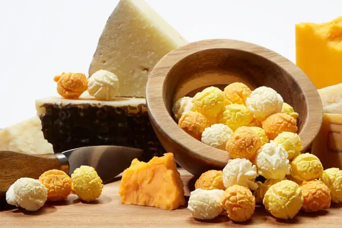 Blocks of cheese next to a bowl of gourmet popcorn kernels flavored Triple Cheese spilled over on a wooden table.