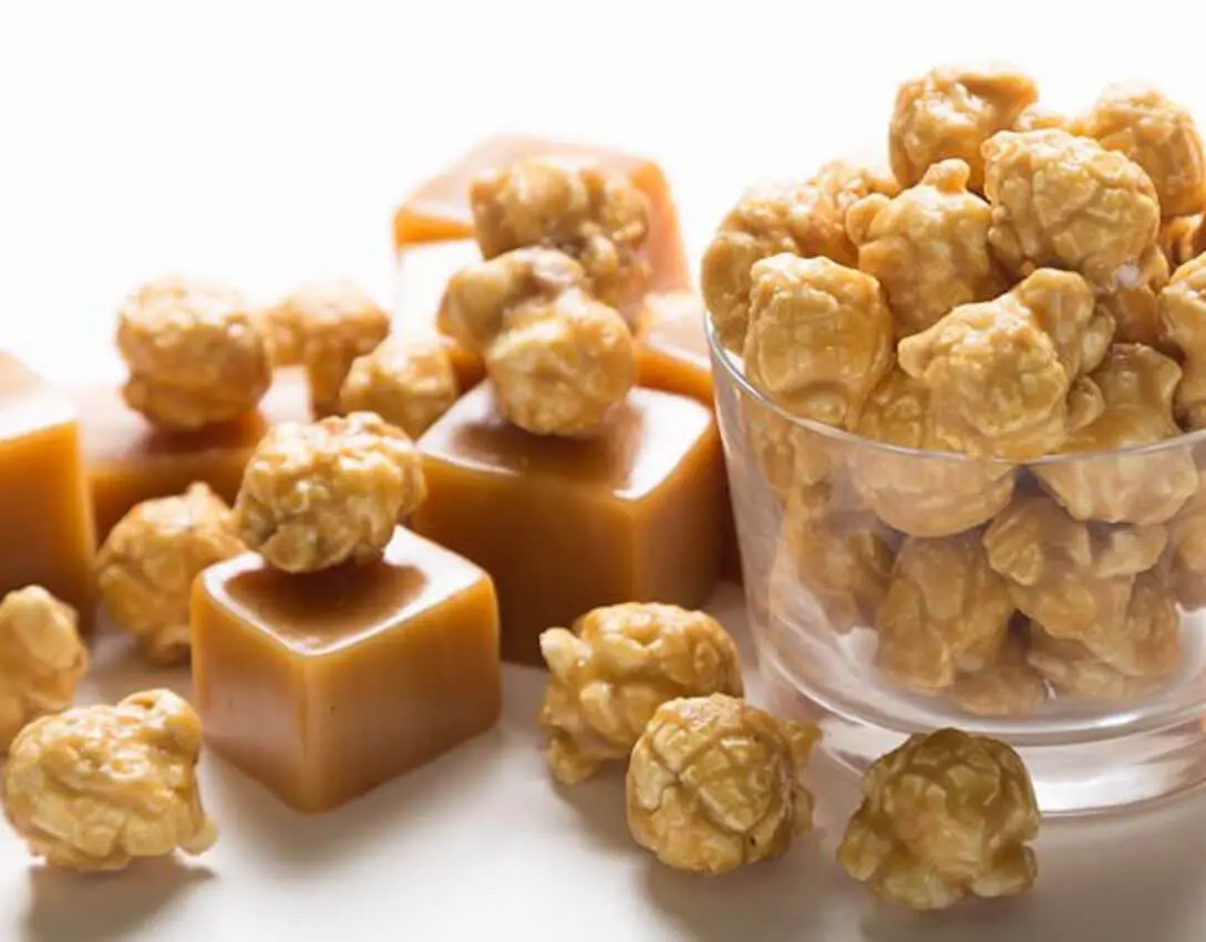 A picture of gourmet popcorn flavored Caramel Popcorn in glass bowl and scattered on white table with cubes of caramel.