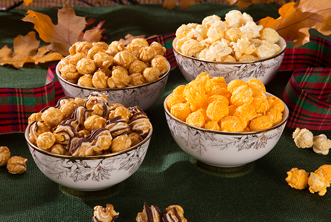 A picture of gourmet Popcornopolis popcorn flavored of Caramel Corn, Kettle, Cheese, and Zebra® popcorn in a white China bowl on a table decorated with plaid ribbon and oak leaves. On top of a green tablecloth with scattered kernels.