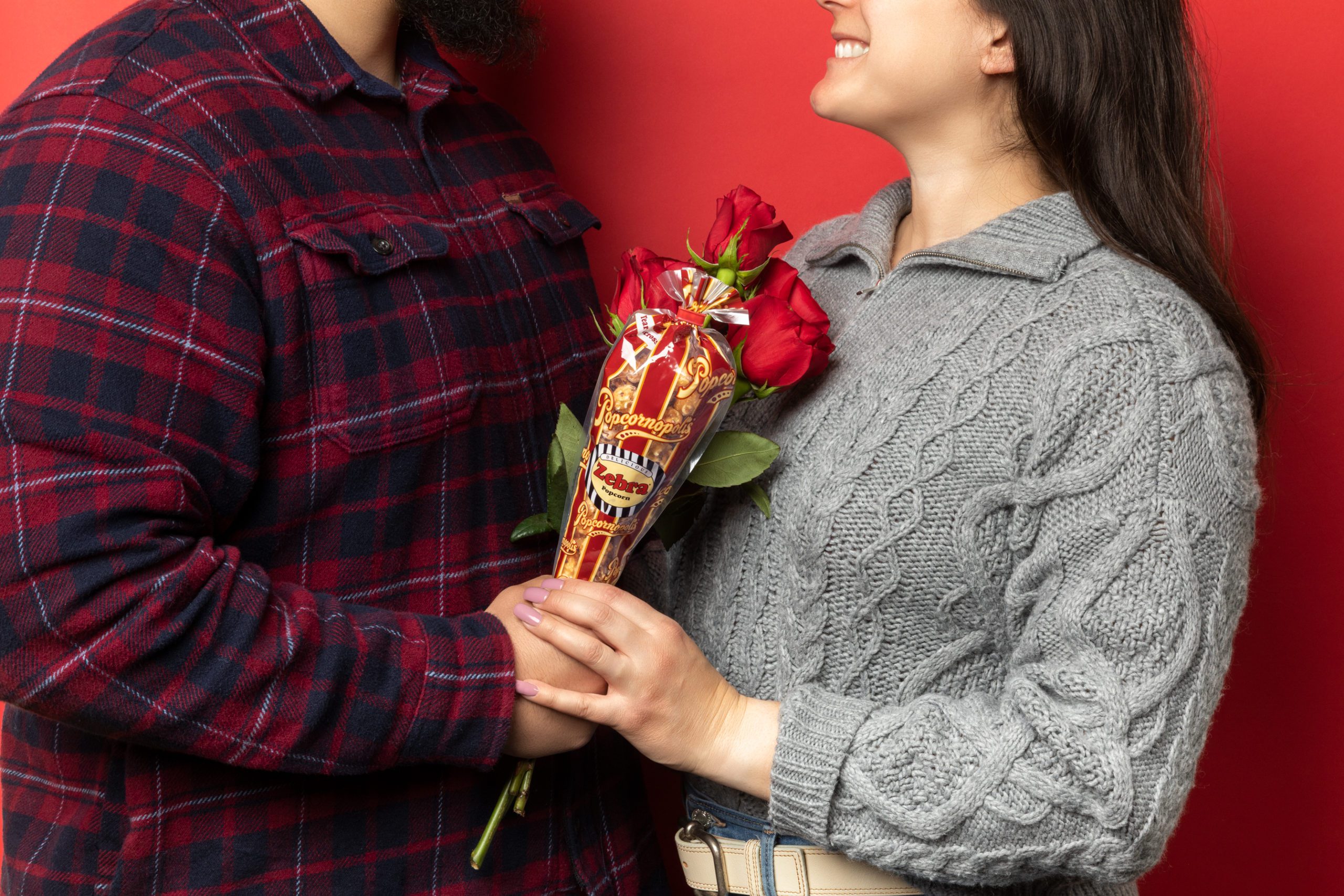 Picture of a man in a stripe red, blue and white flannel holding a mini cone flavored of Zebra® Popcorn and 4 long stem red Rosas. A woman with a gray wool gray sweater holding the man's hand. Red background.